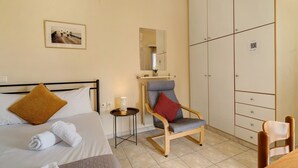A hotel room with a bed, a chair, a small table with a lamp, a wardrobe, and a painting on the wall.