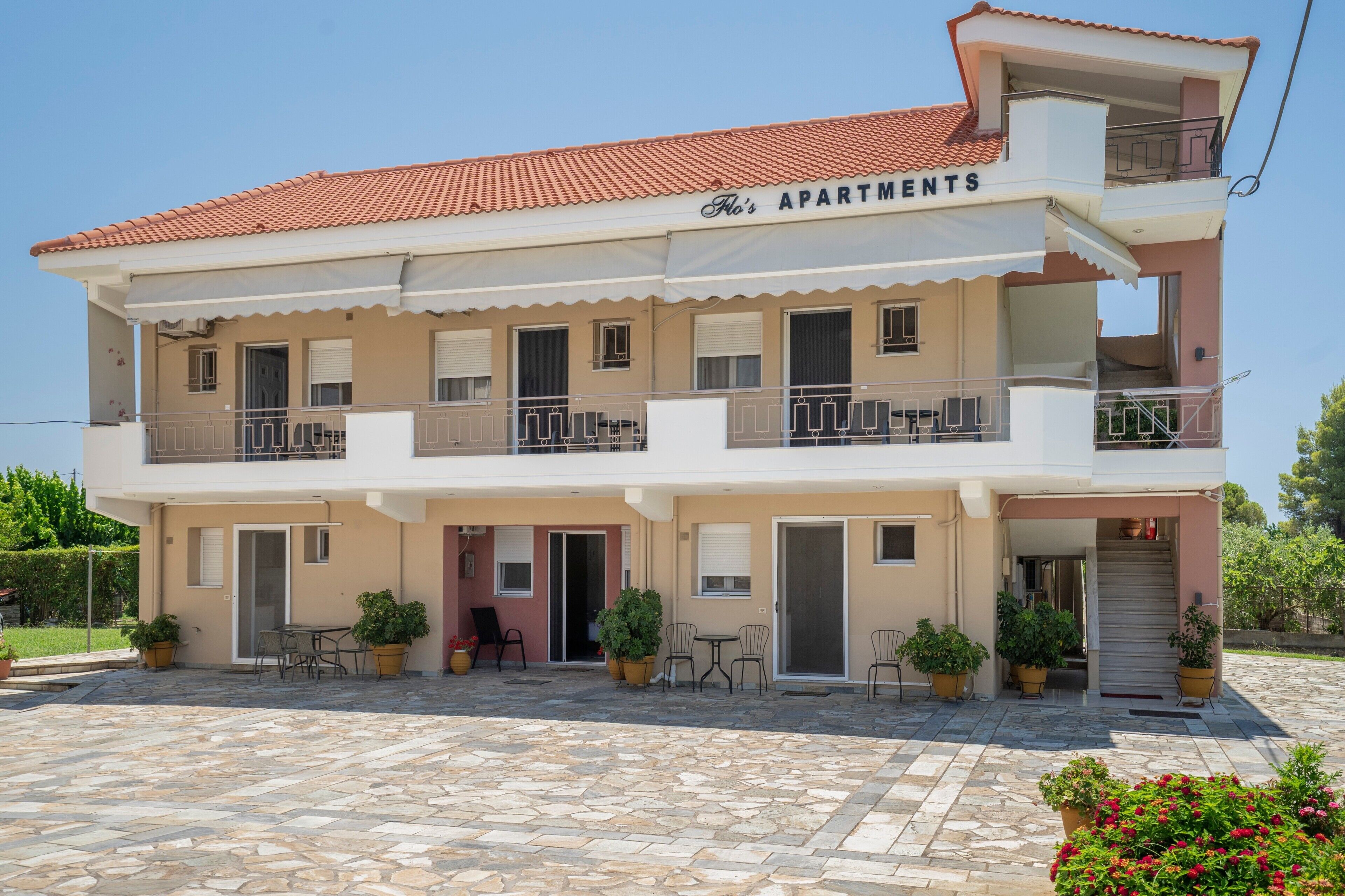 A two-story building with a sign reading 'APARTMENTS' above the entrance. There are multiple balconies with railings, and the ground floor has a paved patio area with outdoor furniture and potted plants.