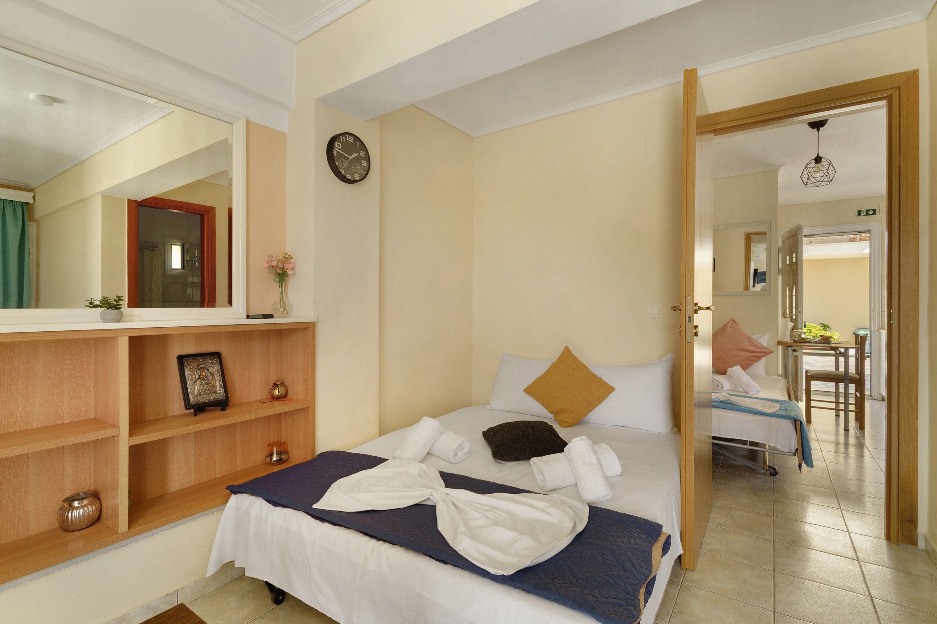 A hotel room with a bed, a bedside table, a mirror, a clock, and a door leading to a bathroom.