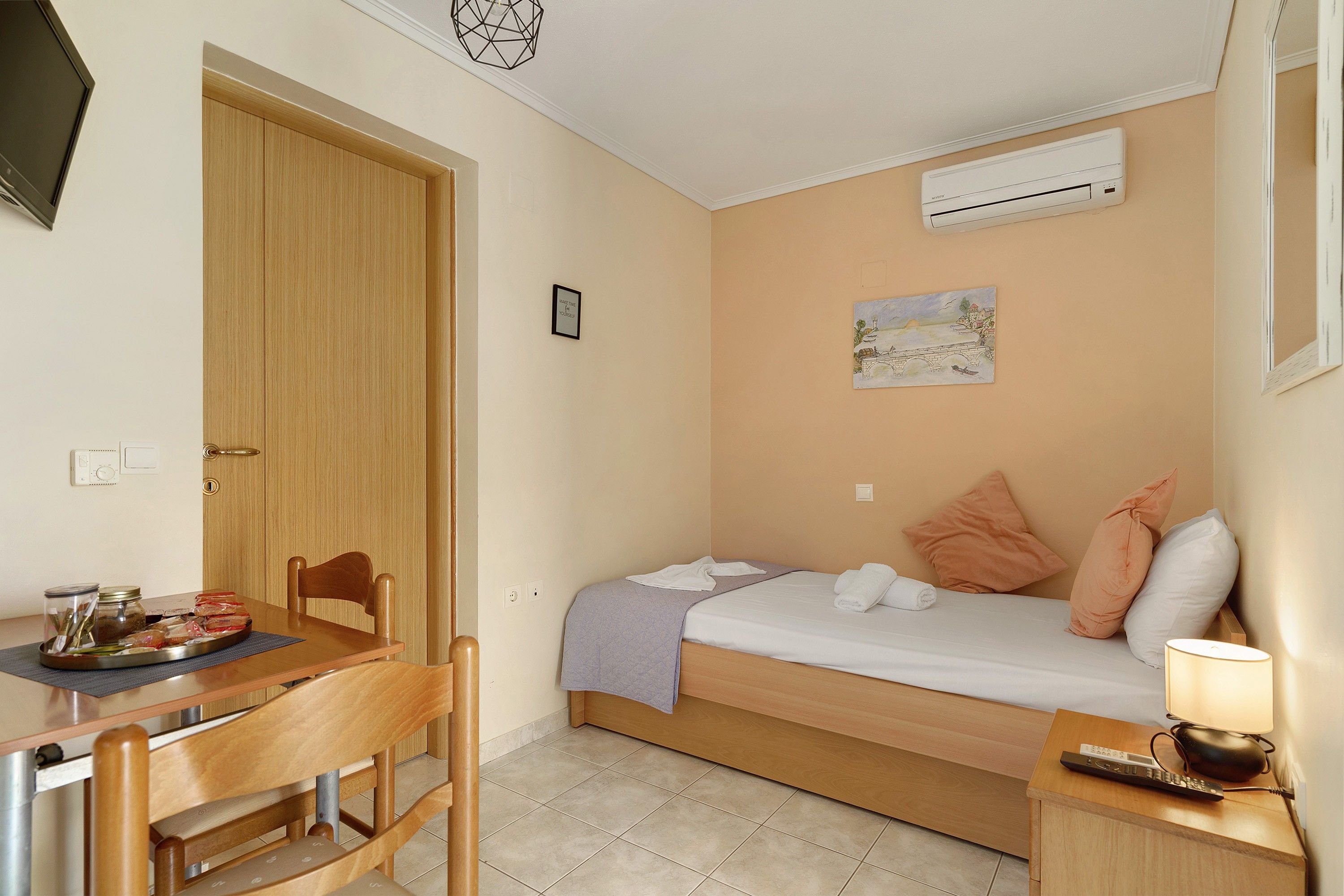 Habitación pequeña con cama, una mesita, una silla, equipo de aire acondicionado y una obra de arte en la pared.