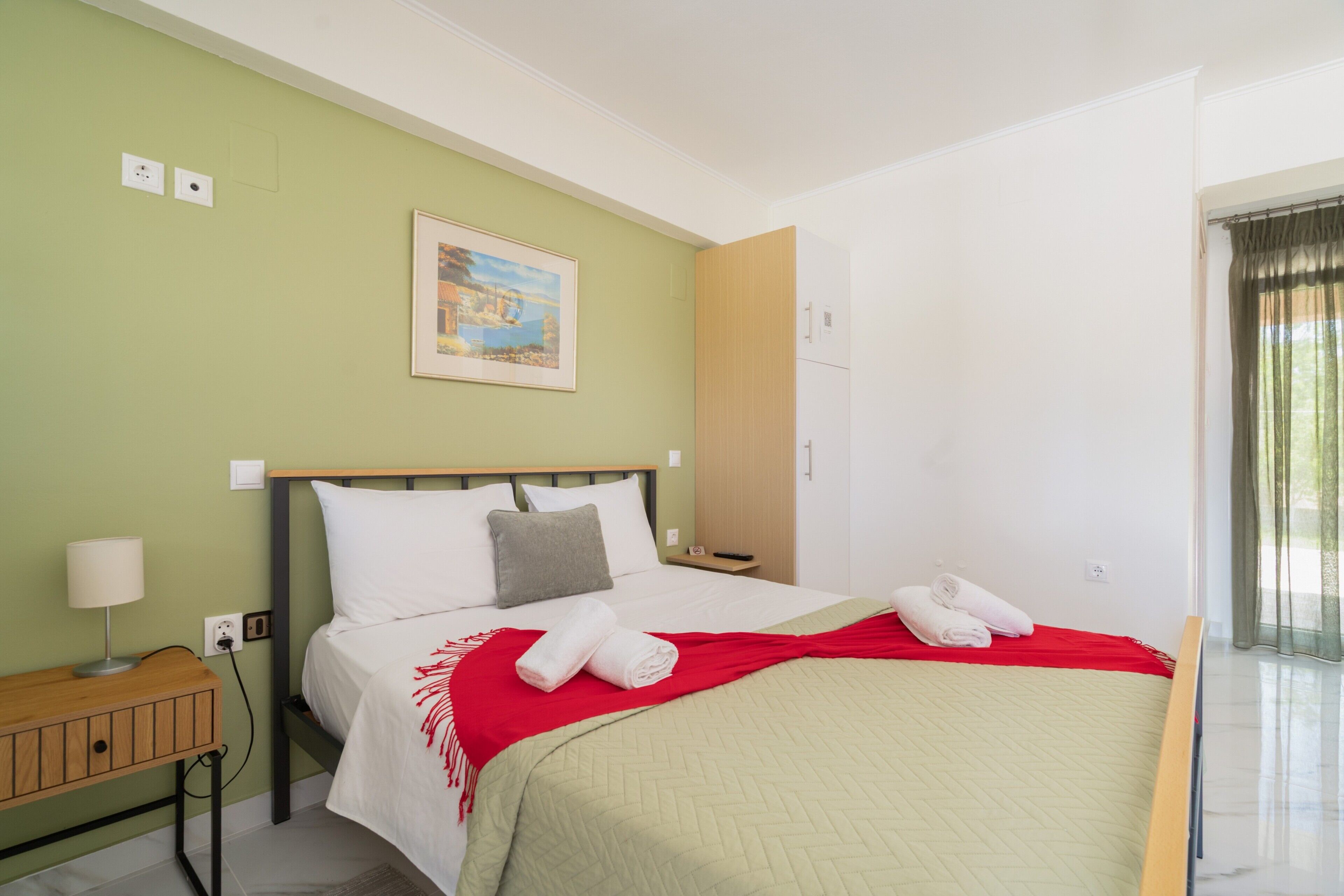 A hotel room with a bed, a nightstand, a lamp, a painting on the wall, and a window with curtains.