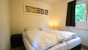 Un dormitorio con cama, lámpara de noche, ventana con cortinas e imagen colgada en la pared.
