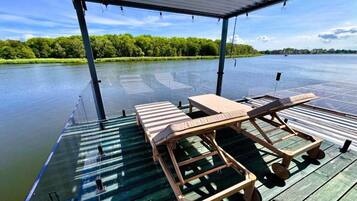 Une terrasse couverte, dotée de deux chaises longues et d’une table, qui offre une vue sur une étendue d’eau calme bordée d’une forêt.