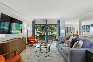 A modern living room with a flat-screen TV, orange and grey sofas, a coffee table, and a view of a garden through large windows. - Golf View Villa Near Harbour Town Marina! (Hilton Head Island)
