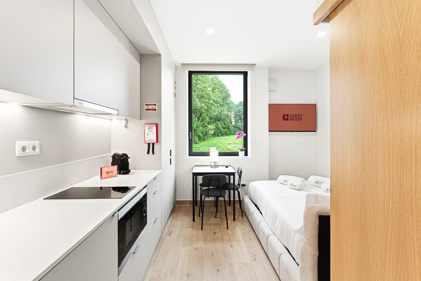 A compact hotel room with a kitchenette, a dining area, a bed, and a window with a view of greenery.