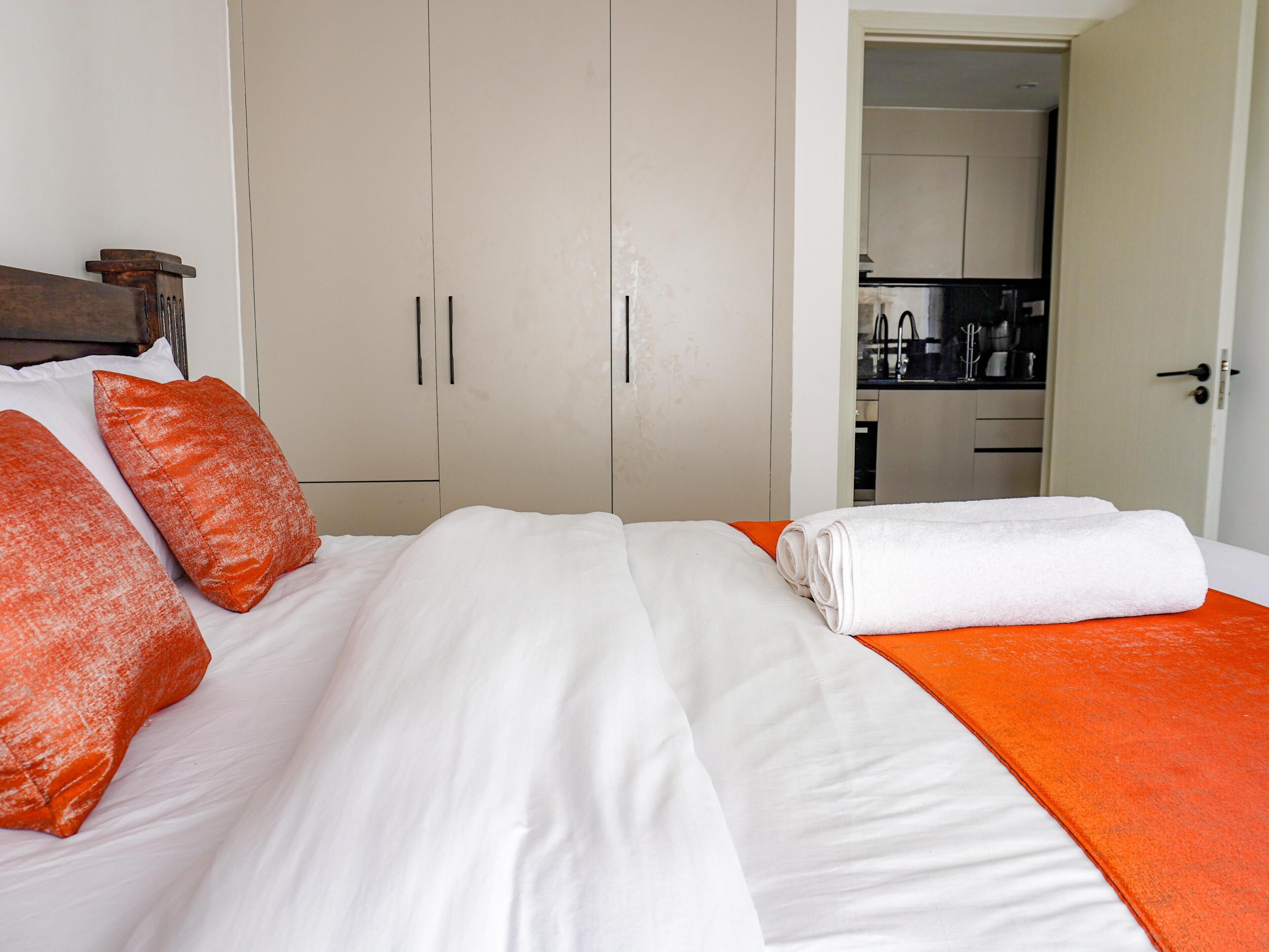 Una habitación de hotel con una cama, un edredón naranja, almohadas blancas y vista a una cocina.