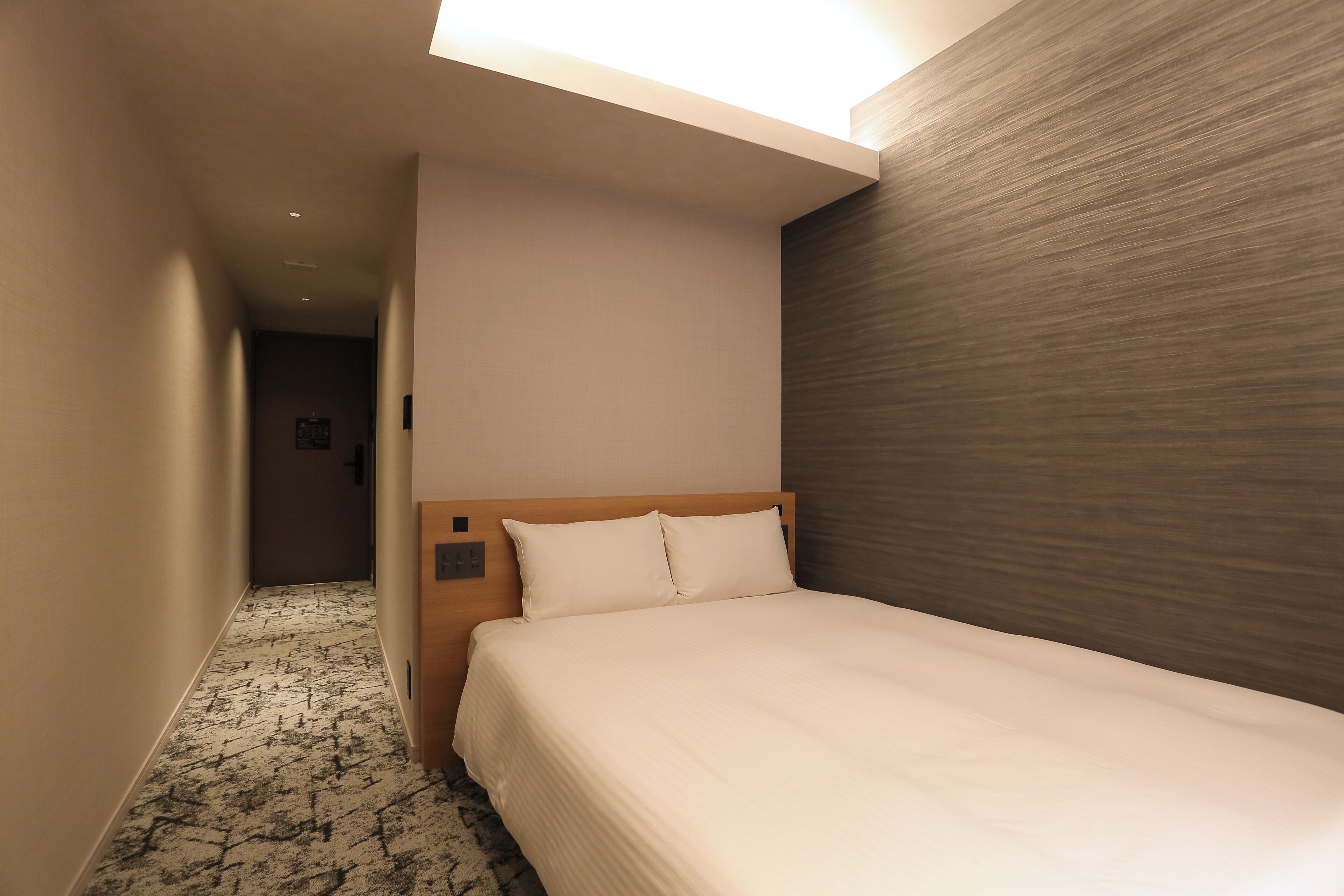 Habitación de hotel con cama, cabecera de madera, piso alfombrado y pared con textura.