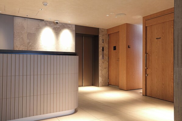 Un vestíbulo de hotel con un mostrador de recepción, una puerta de madera con la etiqueta 'TOILET' y un ascensor.