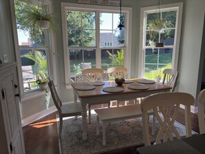 Un comedor con mesa de madera, sillas blancas y vistas a un jardín trasero a través de amplios ventanales.