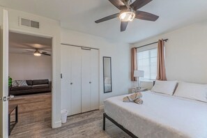 Une chambre à coucher comprenant un lit, un ventilateur de plafond, une fenêtre avec des rideaux, une table de chevet et un miroir.