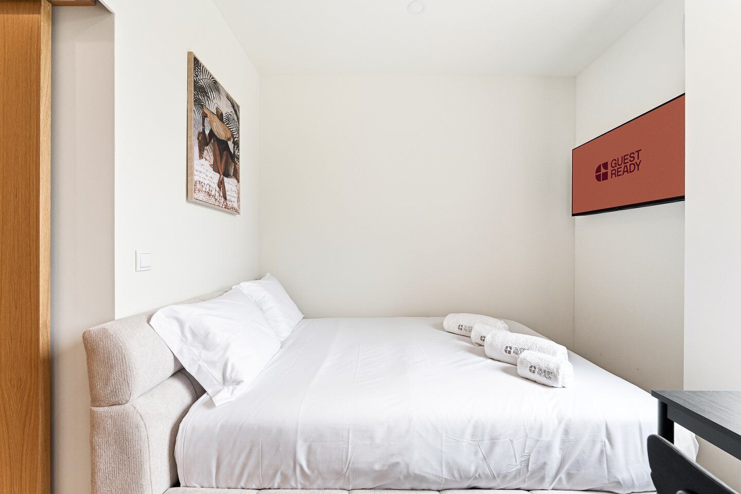 A single bed with white bedding, a small bedside table, a wall-mounted artwork, and a 'GUEST READY' sign.