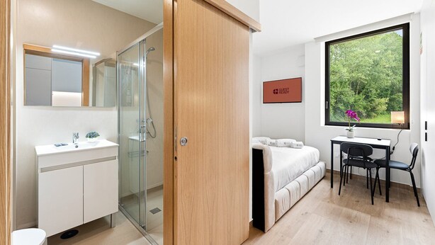 Una moderna camera d'albergo con box doccia in vetro, un vanità bianco, un letto con testiera bianca, una scrivania con sedia e un'ampia finestra con vista sul verde.