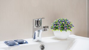 Un lavandino con rubinetto cromato, una pianta in vaso con fiori viola e due confezioni sul lavandino.