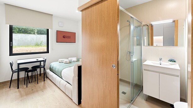 Habitación de hotel moderna con cama, escritorio y sillas, ducha y baño con lavamanos y espejo.