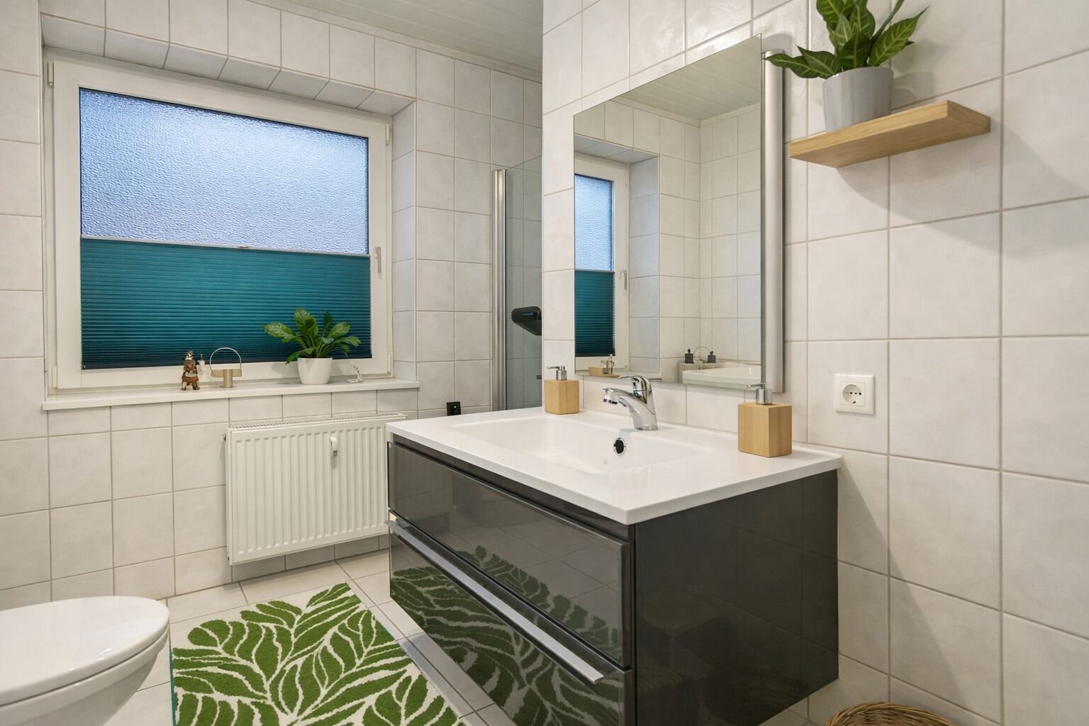 A modern bathroom with a large sink, a mirror, a window with blinds, a radiator, and a green patterned rug.