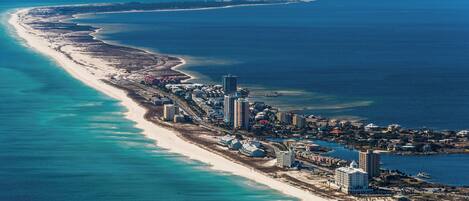 An aerial view of a coastal resort area with a long sandy beach, a pier extending into the ocean, and a cluster of buildings along the shore.