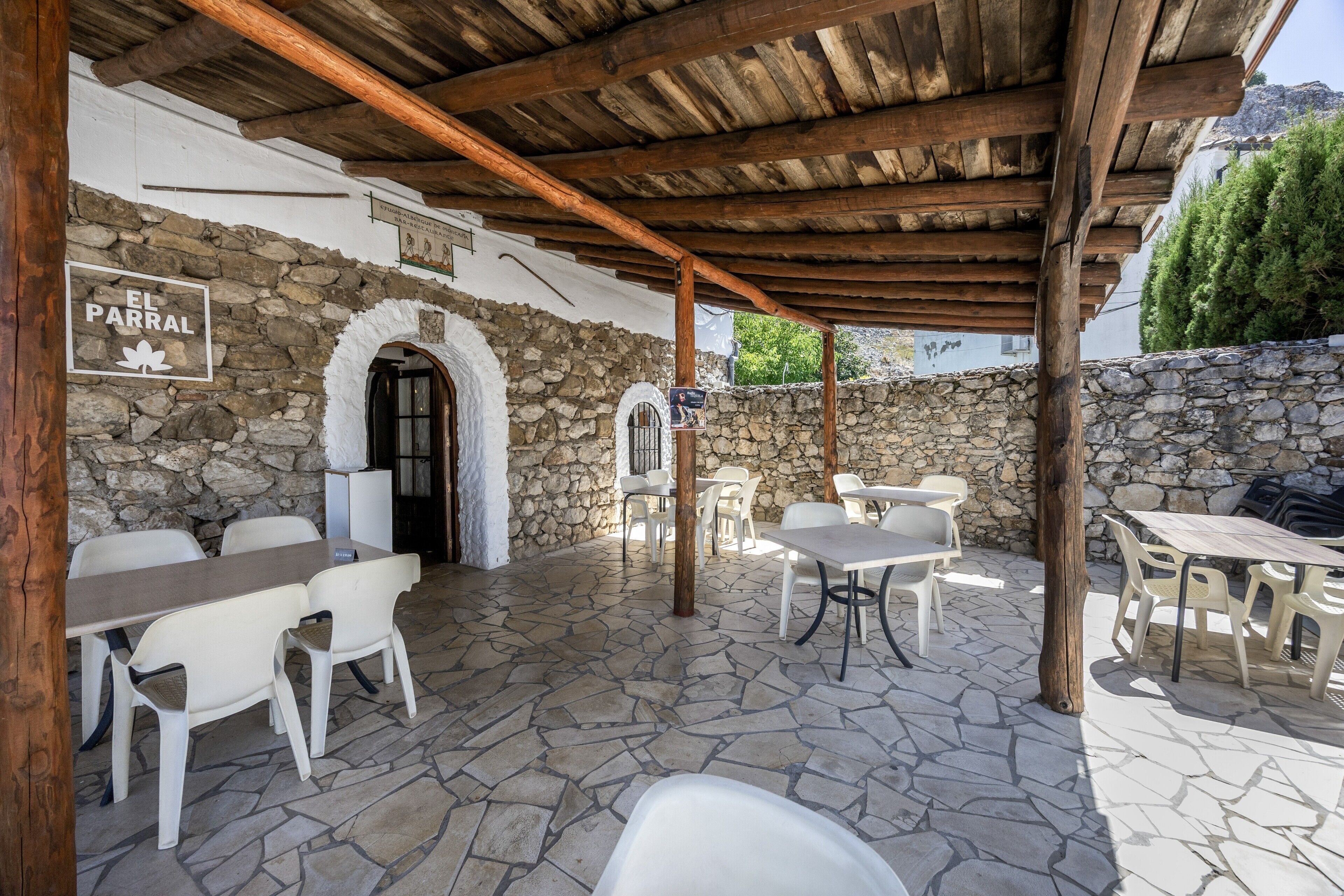Un patio al aire libre con piso de piedra, pérgola de madera, sillas y mesas de plástico blanco.