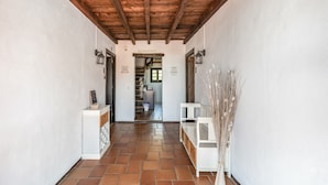 A hallway with a wooden ceiling, white walls, and a tiled floor. There are two white cabinets with a small shelf, a white chair, and a decorative plant.