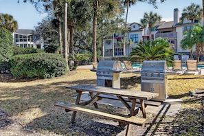 Une table de pique-nique avec deux barbecues, située dans un espace aménagé avec des palmiers et une piscine en arrière-plan.