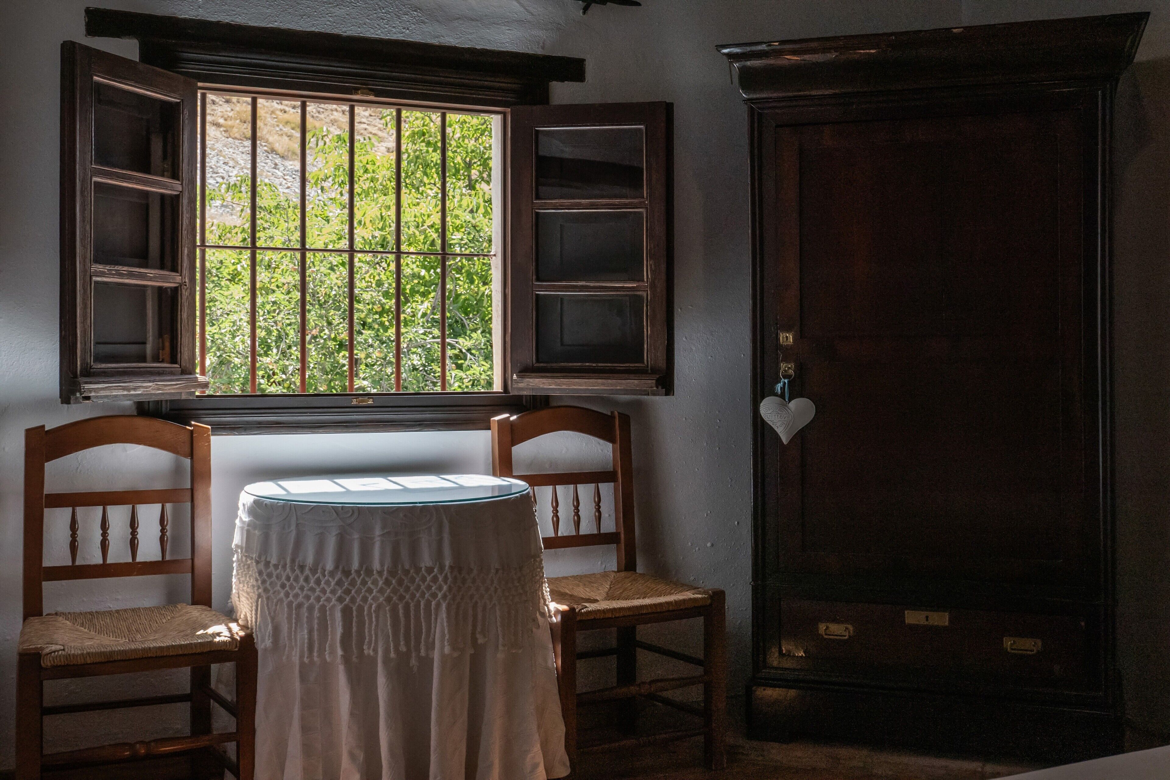 Una habitación con ventana, dos sillas de madera, una mesita redonda y un aparador de madera oscura.