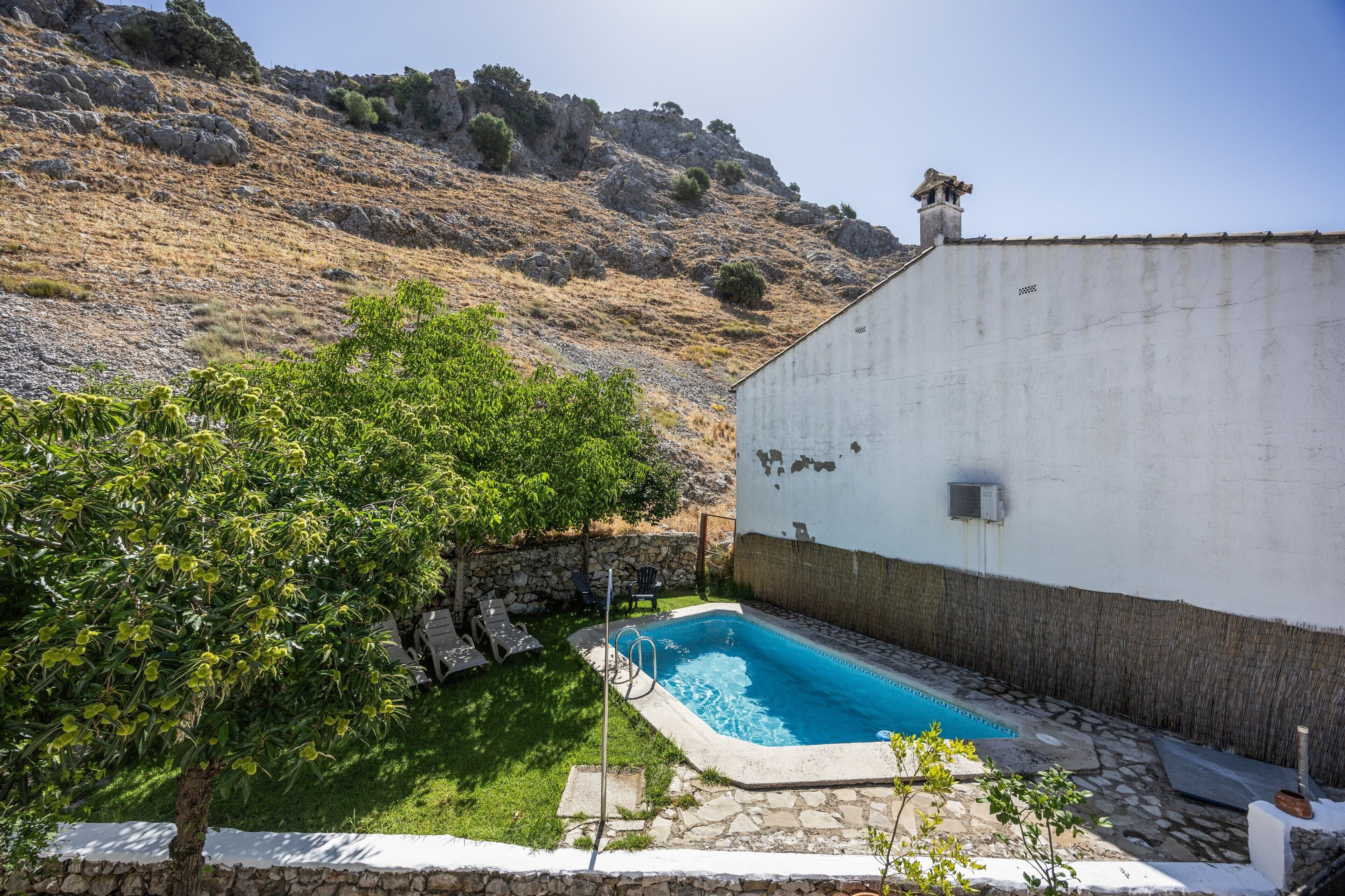 Una piscina con cubierta de piedra, rodeada por un edificio blanco con base de piedra, ubicada frente a una ladera.