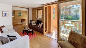 Uma sala de estar aconchegante com um sofá, uma mesa de centro e piso de madeira. Há uma porta de vidro deslizante que dá para uma área externa com uma mesa e cadeiras.