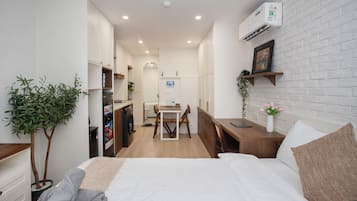 Habitación de hotel pequeña con cama, escritorio, silla, mesita, planta y aire acondicionado de pared.