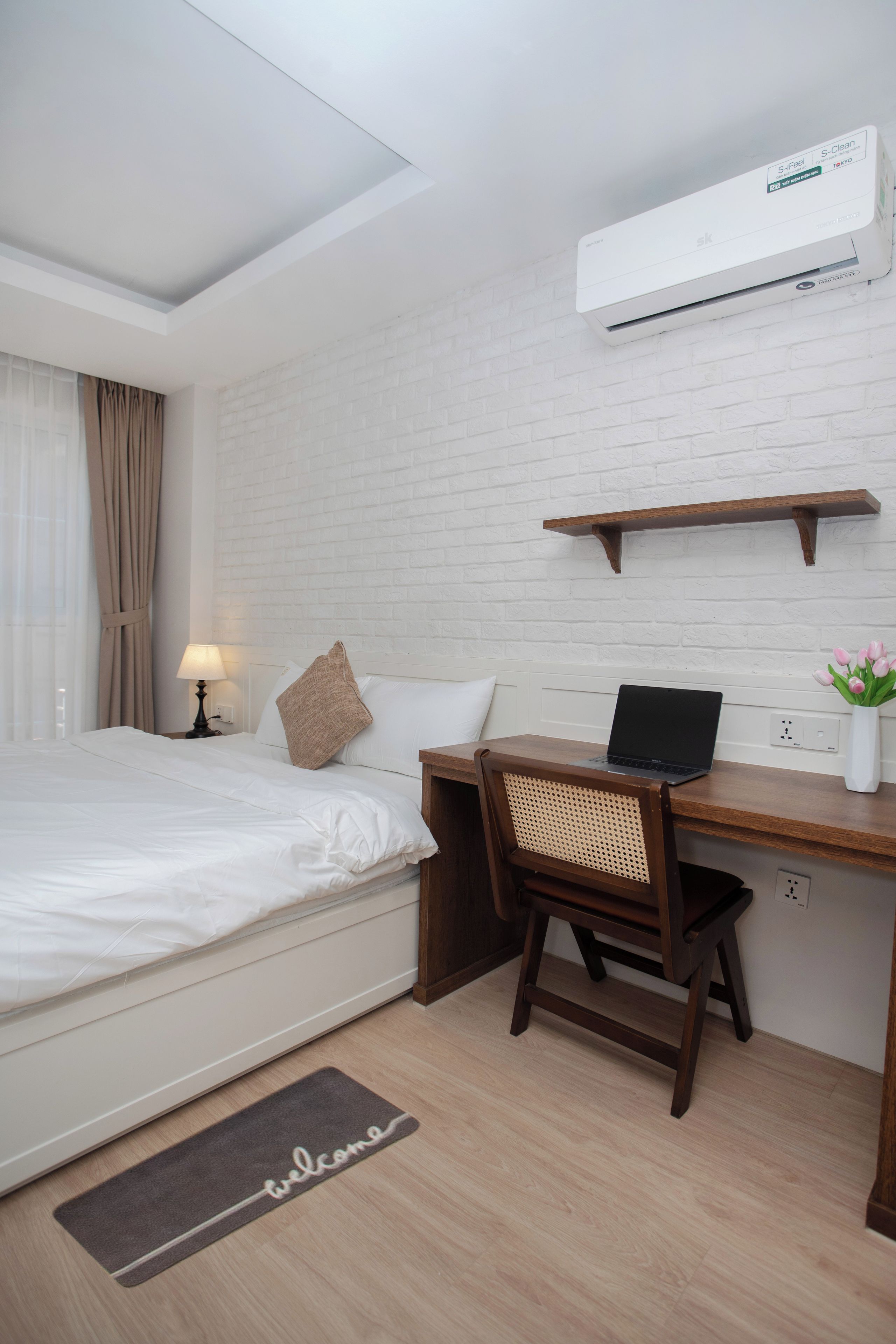 A hotel room with a bed, a desk with a laptop, a chair, an air conditioner, a shelf, and a welcome mat.