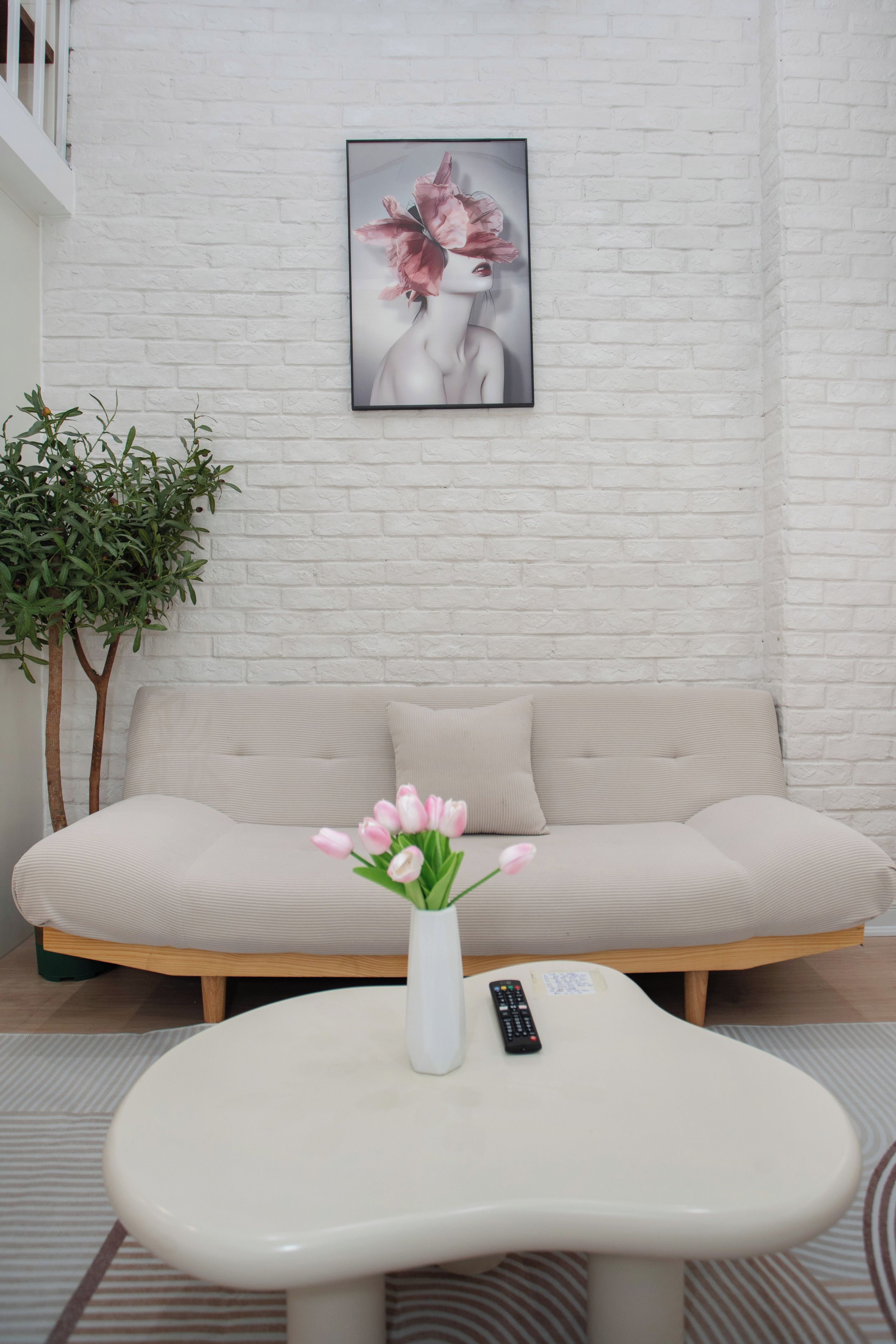 A living room with a white sofa, a white coffee table with a vase of flowers, a remote control, and a potted plant.