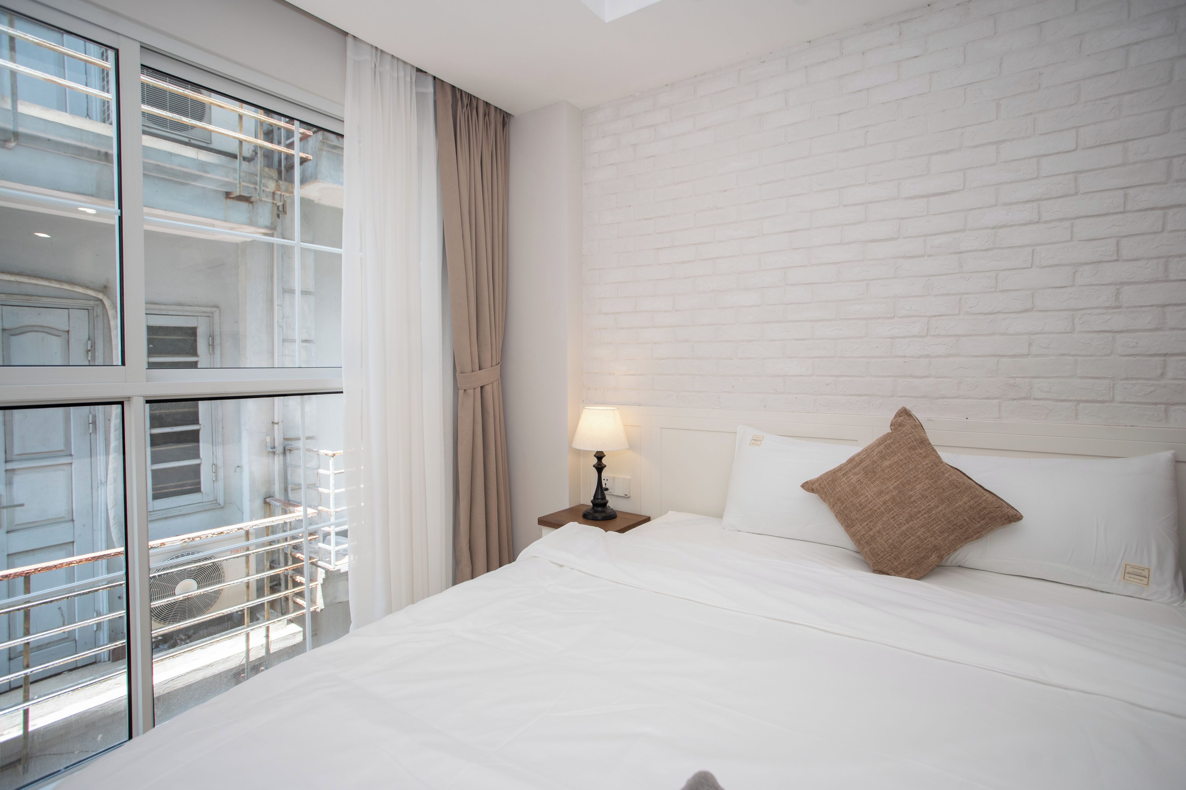 A hotel room with a large window, a bed with white linens, a brown pillow, and a nightstand with a lamp.