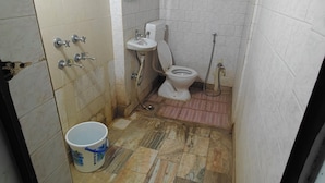 A small, narrow bathroom with a toilet, sink, and shower. The floor is tiled, and there is a bucket on the floor.