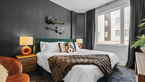 Una camera d'albergo moderna con un letto grande, due comodini, una chitarra appesa al muro e un'ampia finestra con tende.