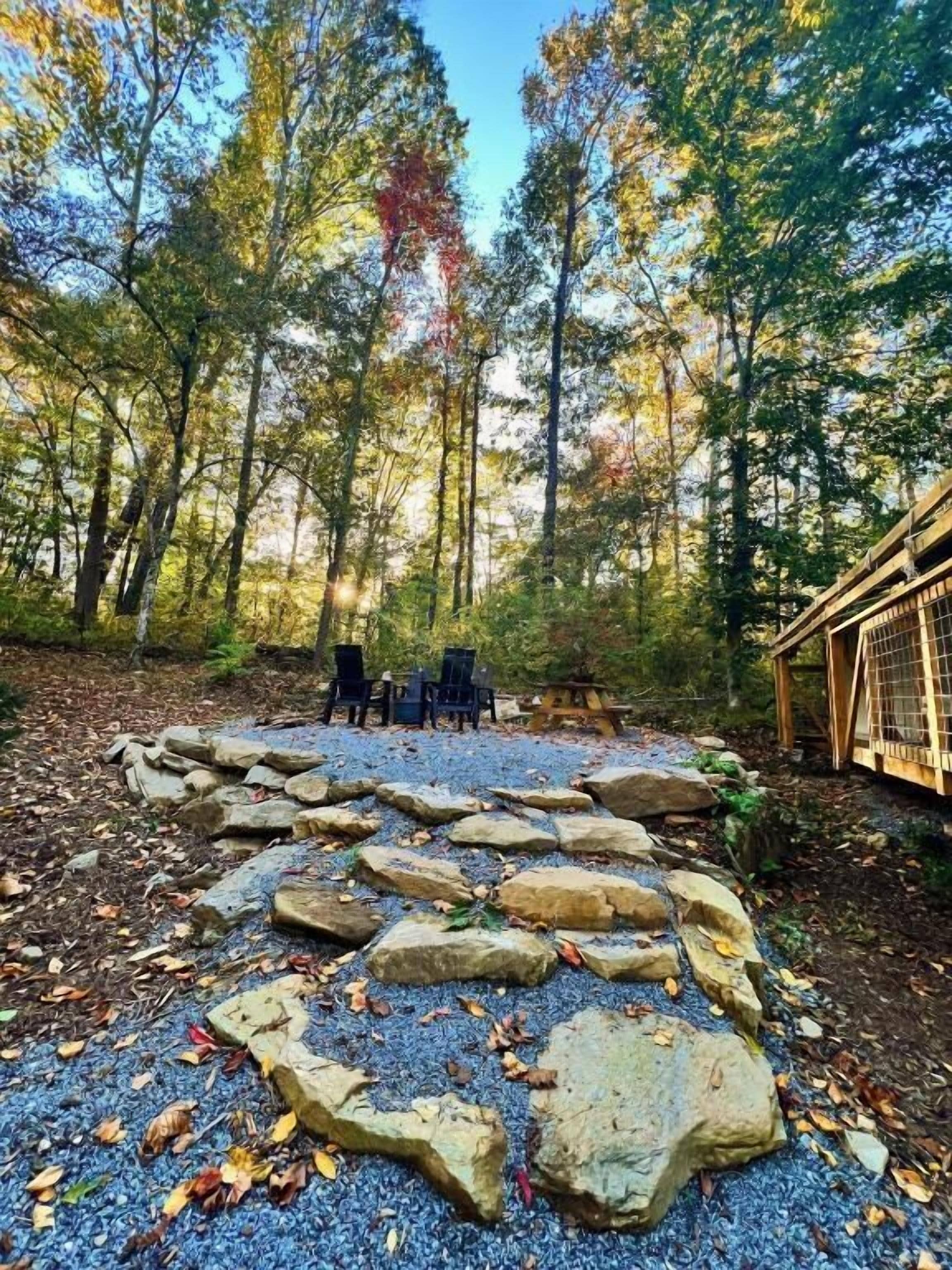 A forested area with a stone pathway leading to a seating area with chairs and a table.