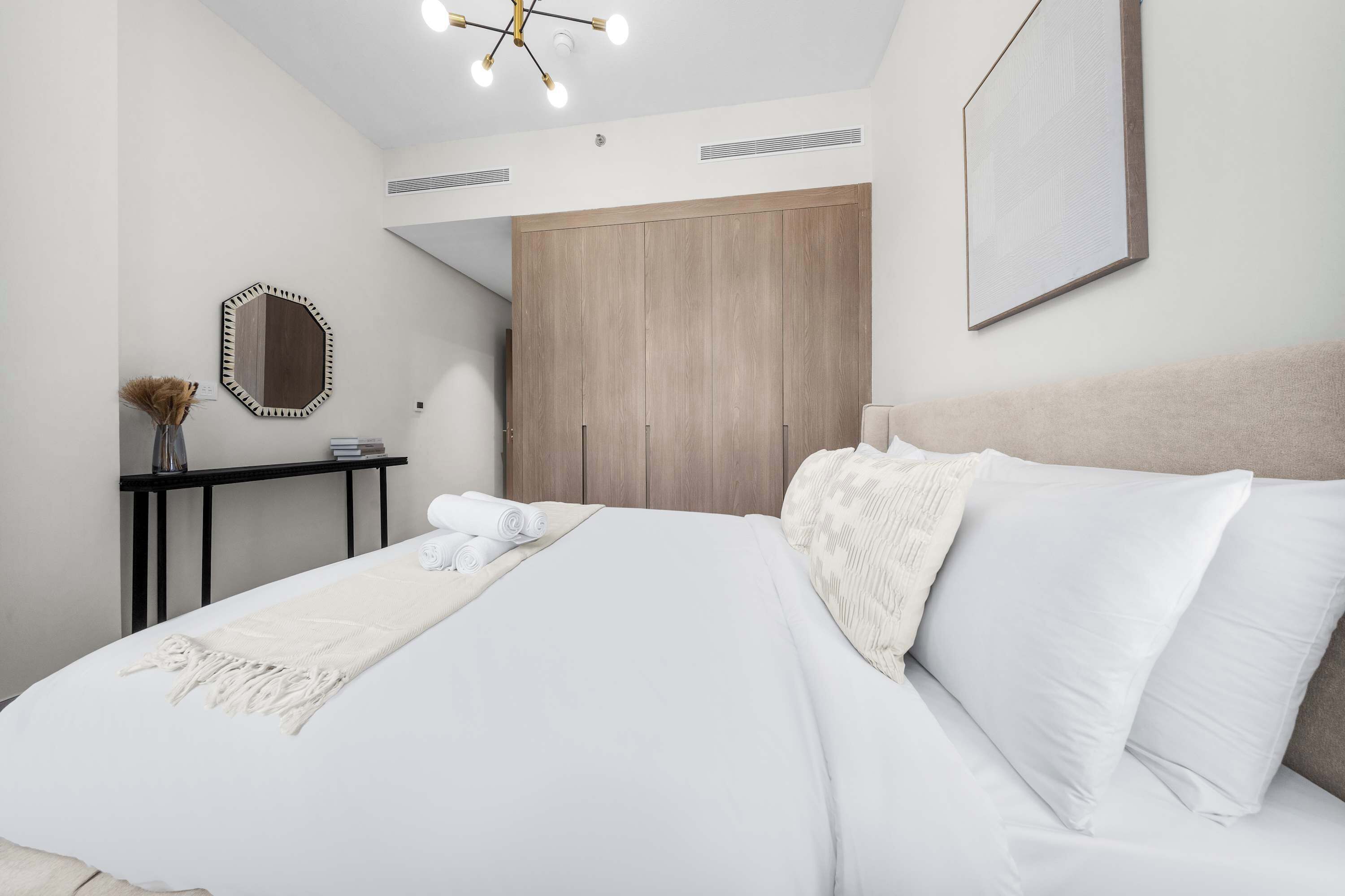 A modern bedroom with a large bed, a wooden wardrobe, a bedside table with a vase, and a ceiling light fixture.