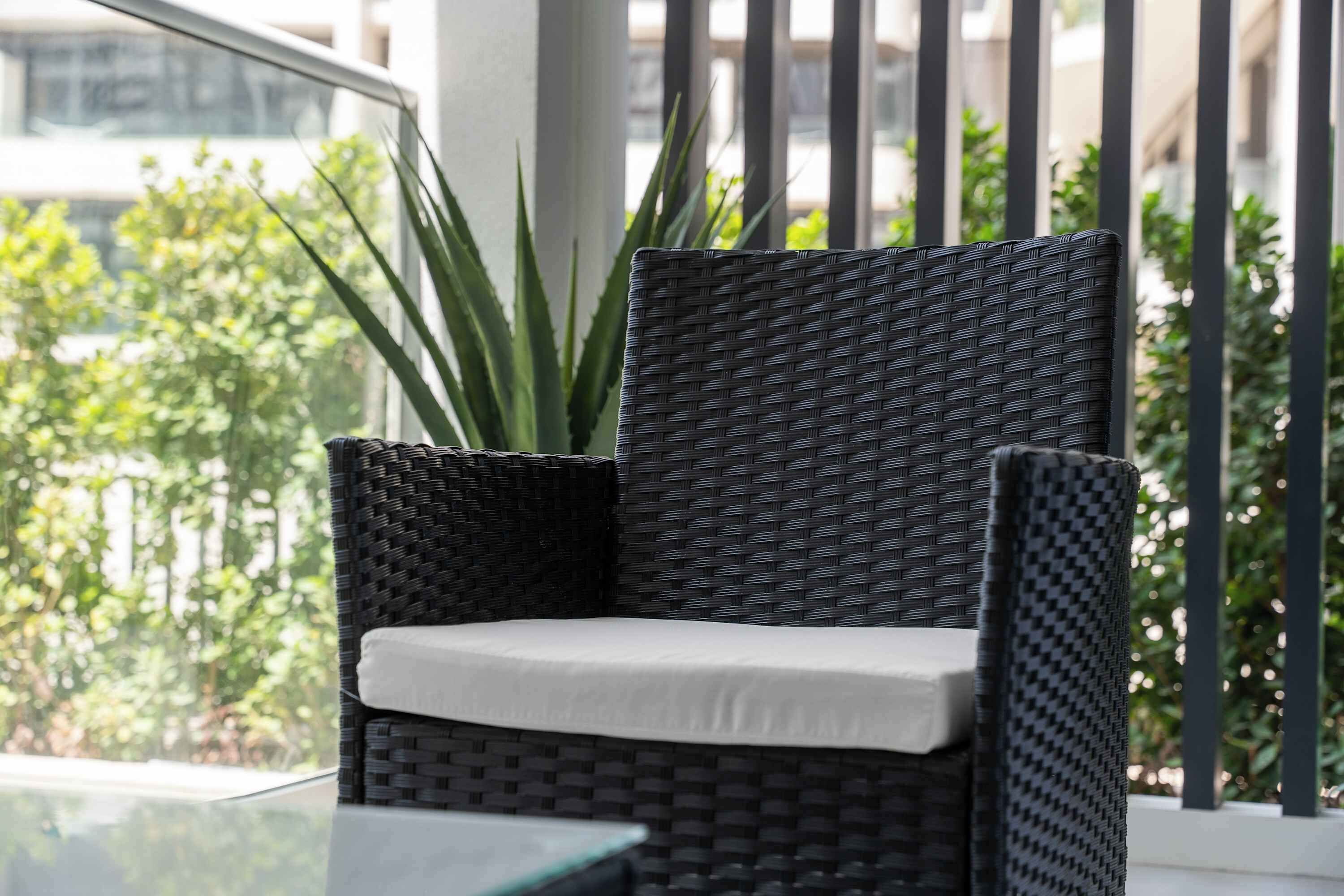 A wicker chair with a cushion, positioned near a glass table and a potted plant, with a view of greenery and a building in the background.