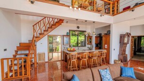 A spacious living area with a wooden staircase, a kitchen with a central island, and a dining area with bar stools. - Casa Infinity | Hilltop Ocean-View | 42' Infinity Pool | Near Manuel Antonio (Portalon)
