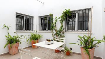 A small patio area with a white table and chairs, surrounded by potted plants and large windows.