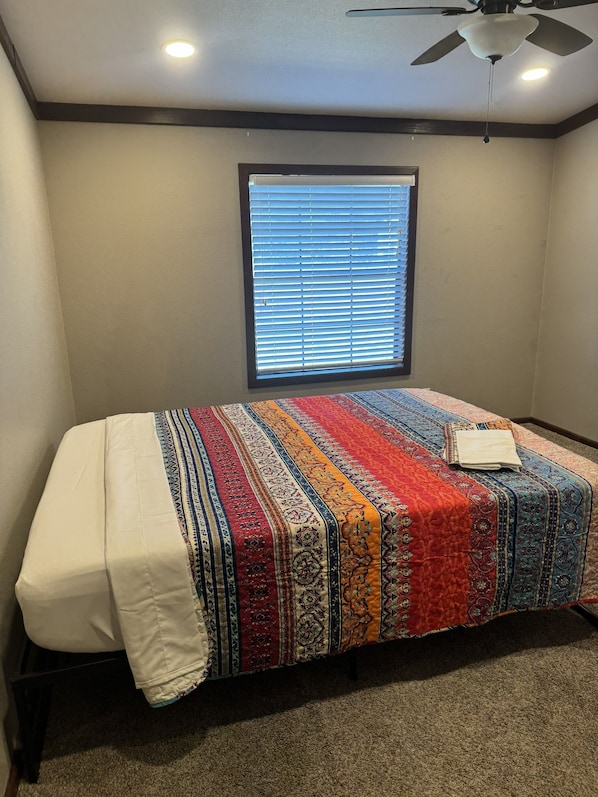 Un dormitorio con cama, ventilador de techo, ventana con persianas y suelo alfombrado.