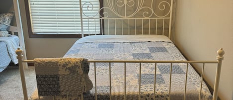 Un lit simple avec un cadre en métal, une fenêtre avec des stores et un sol recouvert de moquette.