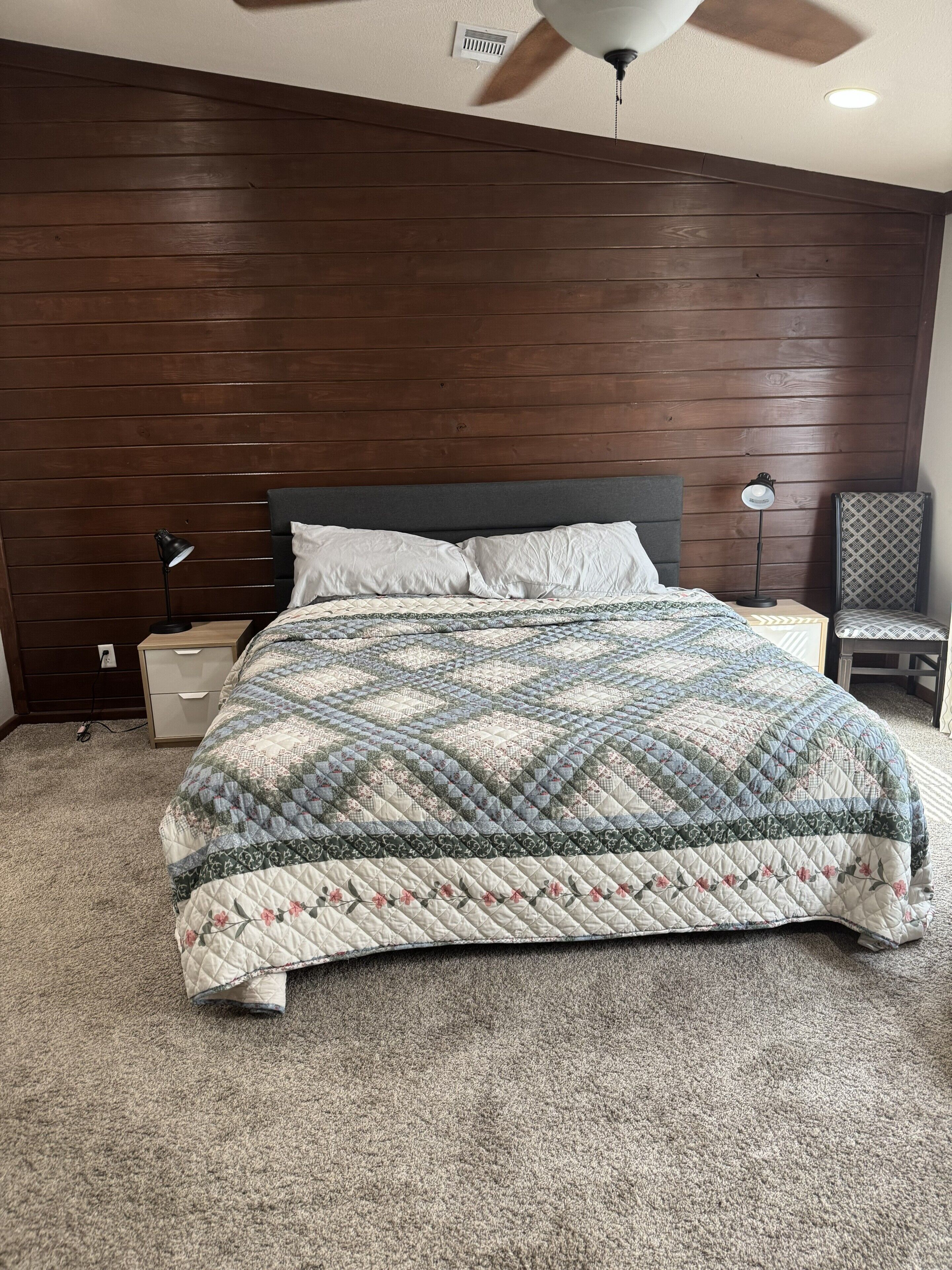 A bedroom with a wooden headboard, a quilted bedspread, a bedside lamp, and a chair.