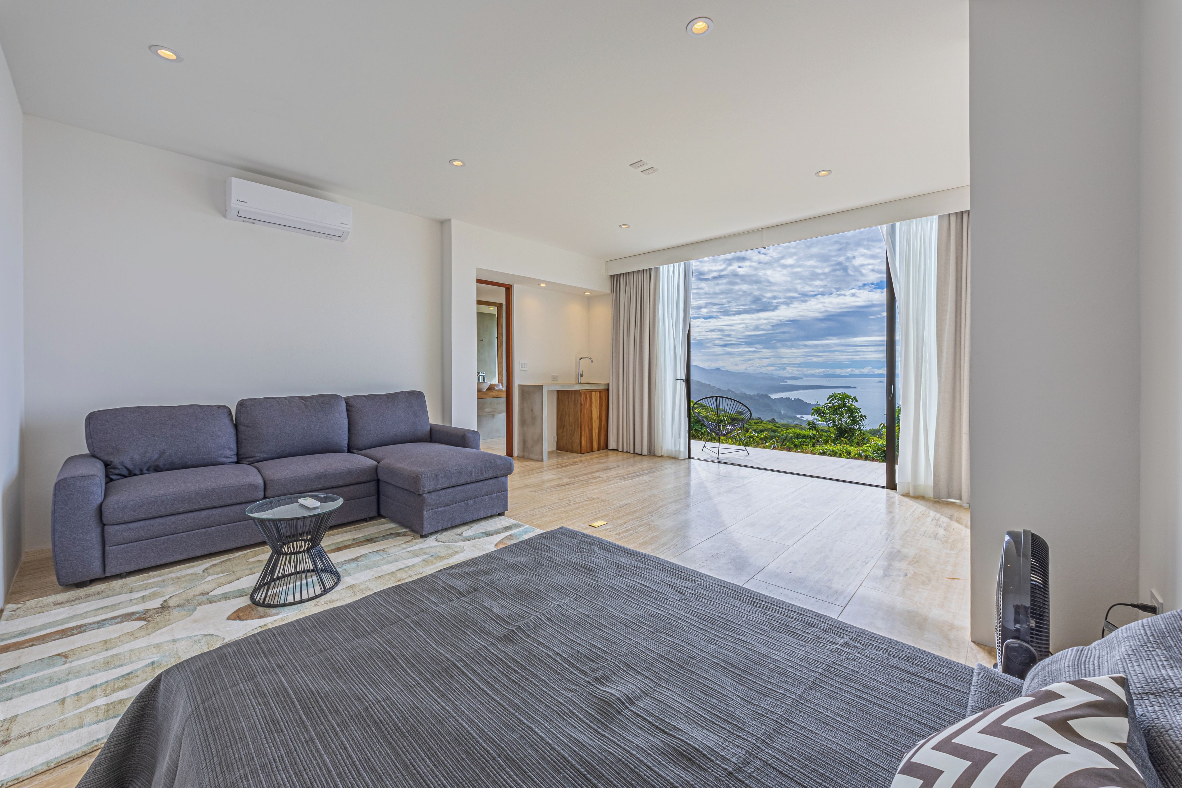 A modern bedroom with a large bed, a grey sofa, and a view of the outdoors.