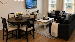 Un salon avec un canapé foncé, une table basse, un téléviseur fixé au mur, une table à manger avec des chaises et un lustre.