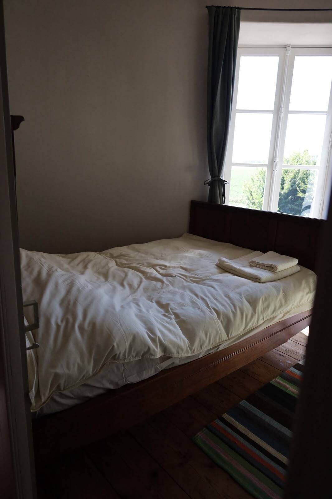 A single bed with a wooden headboard, a window with curtains, and a striped rug on the floor.