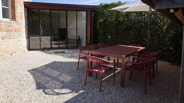A patio area with a table and chairs, a glass door entrance, and a hedge in the background.