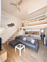 Un apartamento moderno con sofá gris, una mesa de centro blanca, escalera de madera, zona de cocina y ventilador de techo.
