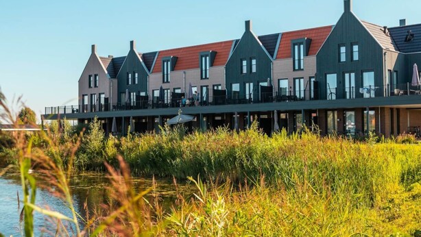 A row of modern, multi-story buildings with a flat roof, connected by a covered walkway, overlooking a body of water. - Reevemeerzicht 6 (Kampen)