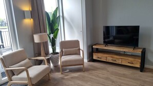 A modern living room with two chairs, a TV on a wooden stand, a potted plant, and a large window. - Reevemeerzicht 6 (Kampen)