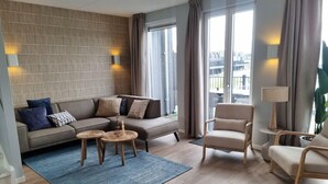 A modern living room with a sofa, two side tables, a chair, and a large window with curtains. - Reevemeerzicht 6 (Kampen)