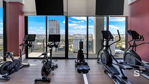 A modern gym with various cardio machines, large windows offering city views, and mounted TVs. - 28th-Floor Condo in Miami Worldcenter (Miami)