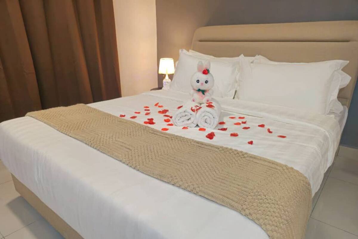 Una cama bien hecha con una toalla enrollada, un peluche y pétalos de rosa sobre la cama.